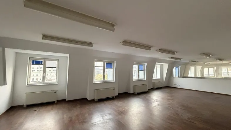Geräumiger, leerer Raum mit dunklem Holzboden, weißen Wänden und mehreren Fenstern mit Stadtblick.