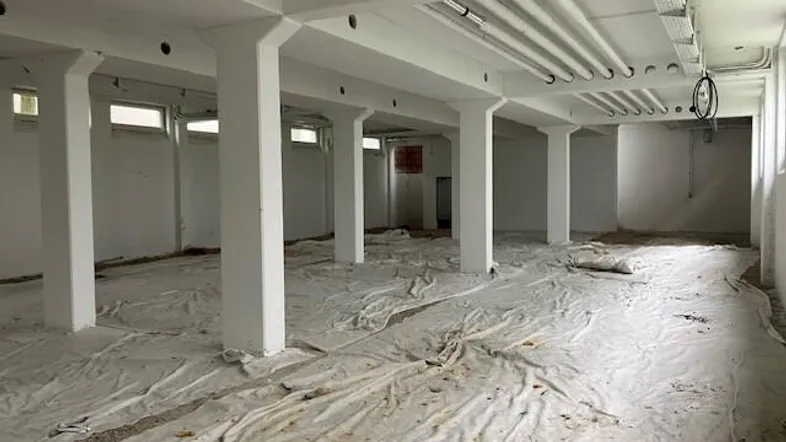 Großer, leerer Raum mit vielen Säulen, sichtbaren Rohren an der Decke und kleinen Fenstern.