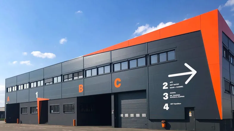 Modernes Industriegebäude mit dunkelgrauer Fassade, orangefarbenen Akzenten und großen Rolltoren unter blauem Himmel.