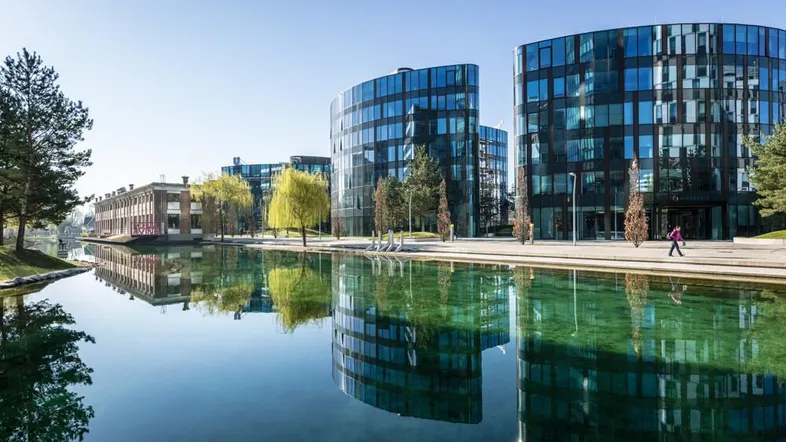 Moderne Bürogebäude mit Glasfassaden spiegeln sich in einem ruhigen Wasserbecken unter blauem Himmel wider.