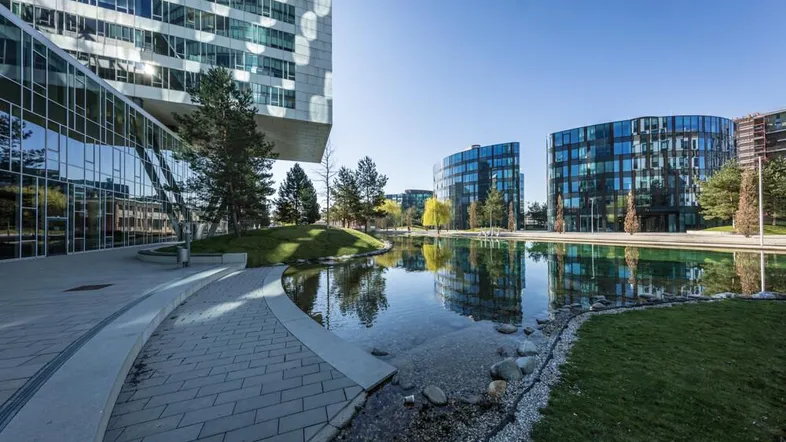 Moderne Bürogebäude mit Glasfassaden spiegeln sich in einem Teich wider, umgeben von grünen Landschaften.