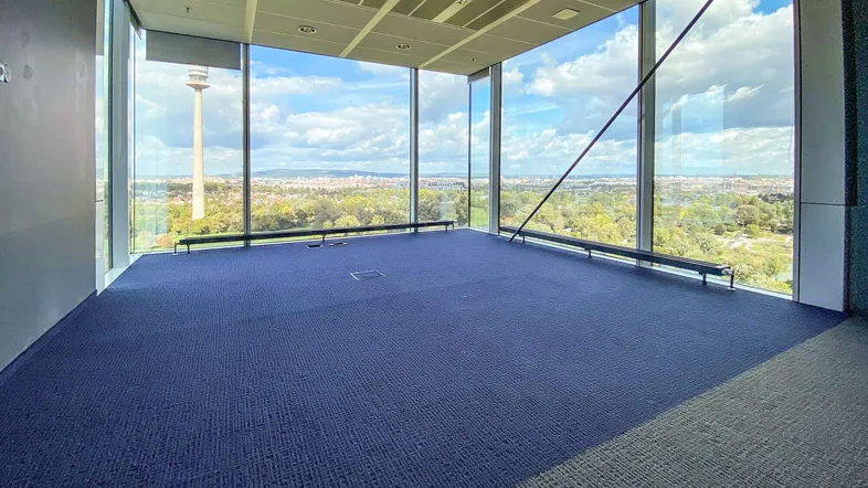 Geräumiges Büro mit großen Fenstern, blauem Teppichboden und Panoramablick auf die Stadt und den Donauturm.