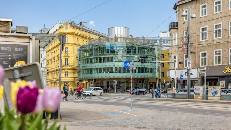 Straßenansicht mit einem modernen Glasgebäude und traditionellen Gebäuden in einer städtischen Umgebung.