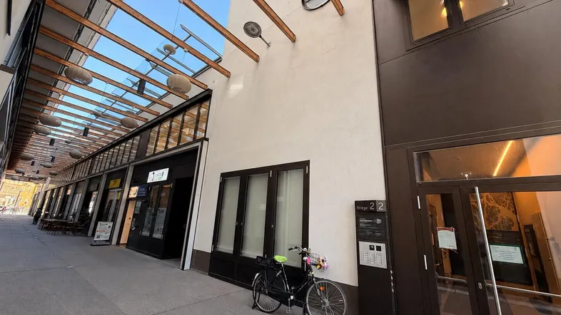 Eingangsbereich eines modernen Gebäudes mit Geschäften und einer Fahrradabstellmöglichkeit unter einem Glasdach.