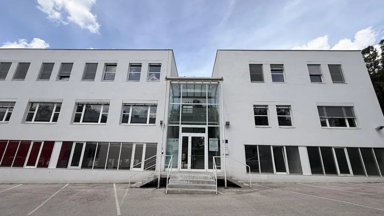Modernes Bürogebäude mit weißer Fassade, vielen Fenstern und einem gläsernen Eingangsbereich unter bewölktem Himmel.