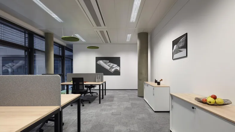Modernes Büro mit Schreibtischen, Trennwänden und großen Fenstern, das viel Tageslicht hereinlässt.
