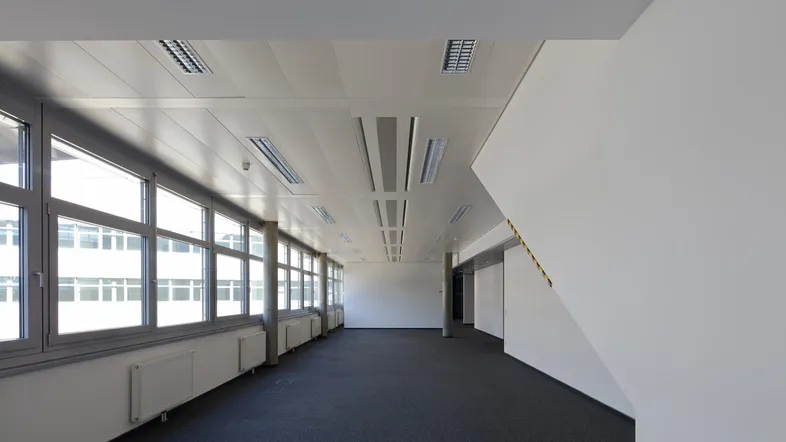 Geräumiges, leeres Büro mit großen Fenstern, dunklem Teppichboden und moderner Deckenbeleuchtung, bereit zur Einrichtung.