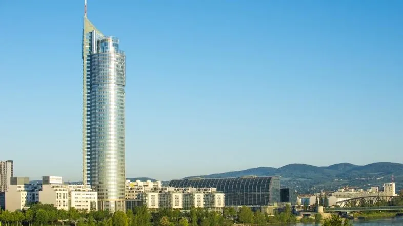 Panoramablick auf ein modernes Hochhaus am Flussufer mit Bergen im Hintergrund bei Tageslicht.