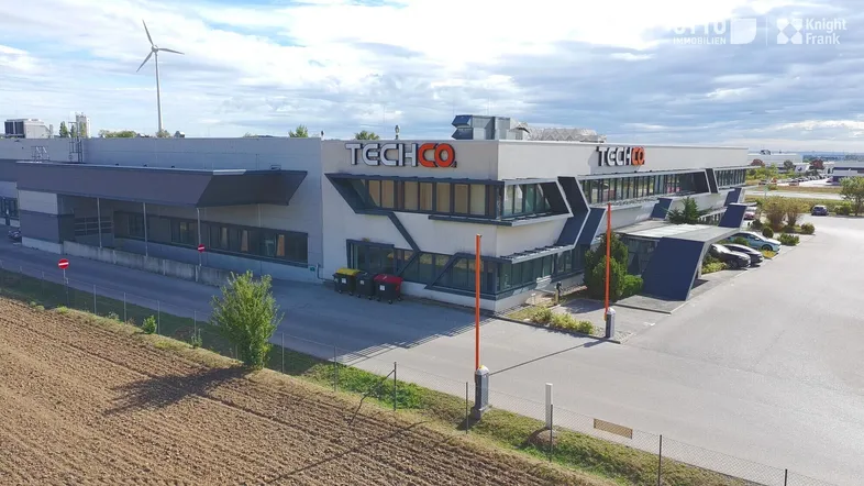 Moderne Gewerbeimmobilie mit Windrad im Hintergrund, Parkplätzen und weitem Blick über die Landschaft.
