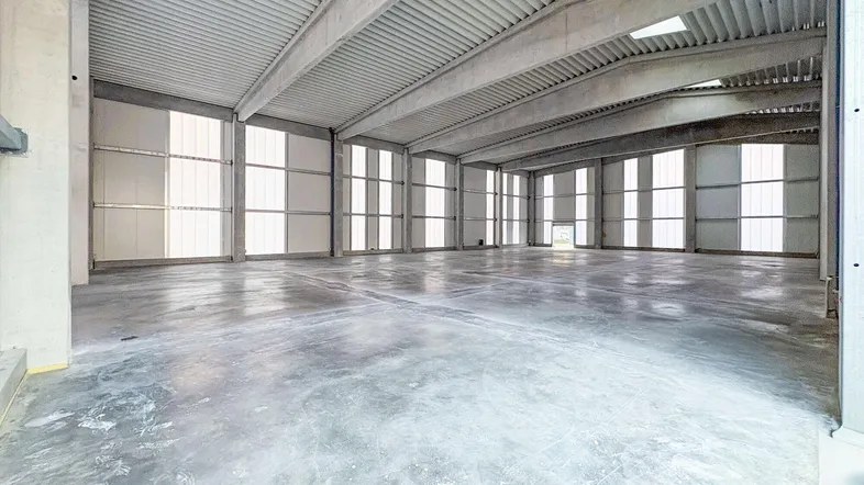 Große, leere Lagerhalle mit Betonboden und Tageslicht durch hohe Fenster und Oberlichter.