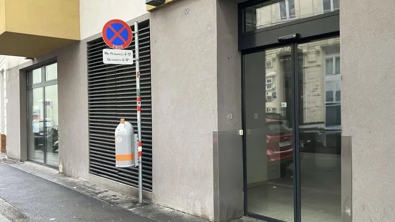 Eingangsbereich eines modernen Gebäudes mit großen Glastüren und einer grauen Fassade von der Straße aus gesehen.