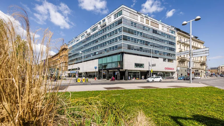 Modernes Bürogebäude mit Glasfassade an einer belebten Straße, umgeben von Grünflächen und blauem Himmel.