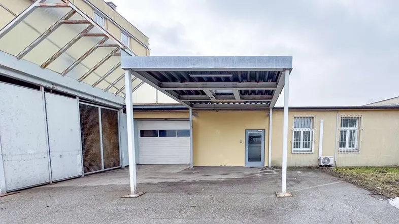 Metall-Carport vor einem Gebäude mit gelber Fassade und Garagentor, umgeben von einer asphaltierten Fläche.