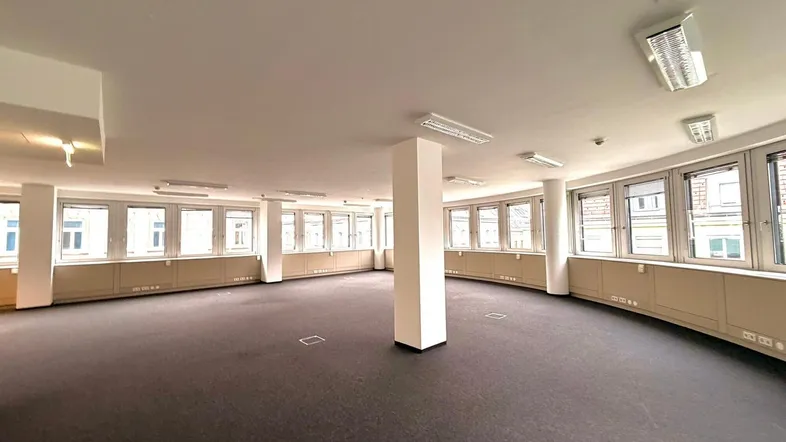 Großer, leerer Büroraum mit Teppichboden, vielen Fenstern und natürlichem Lichteinfall.
