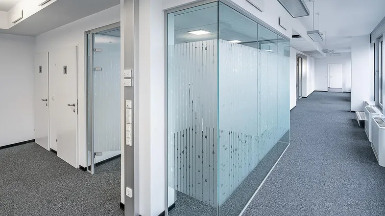 Heller Büroflur mit modernen Glaswänden und Teppichboden, der zu verschiedenen Büroräumen führt.