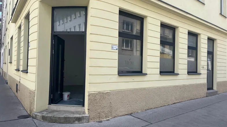 Eckgebäude mit heller Fassade und großen Fenstern, das eine Gewerbefläche im Erdgeschoss zeigt und Renovierungsbedarf andeutet.