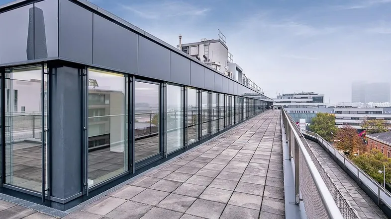 Lange Dachterrasse mit Fliesenboden und Blick auf die Stadt, umgeben von modernen Glasfassaden.
