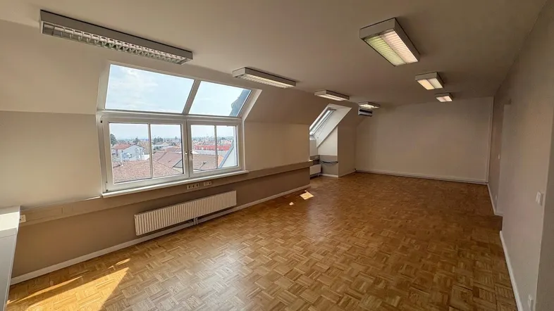 Geräumiger Büroraum im Dachgeschoss mit großen Fenstern, die viel Tageslicht hereinlassen und Stadtblick bieten.