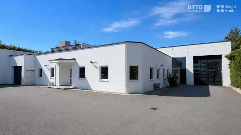 Modernes Geschäftsgebäude mit weißer Fassade und großem asphaltierten Parkplatz unter blauem Himmel.