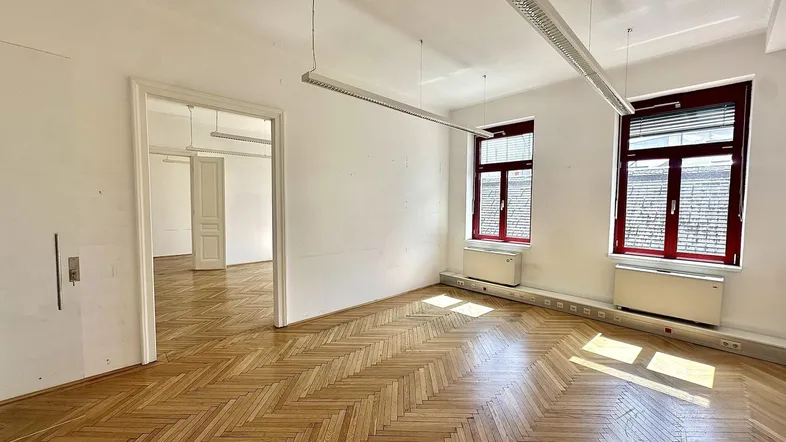 Heller Büroraum mit Fischgrätparkett und großen Fenstern, die viel Tageslicht hereinlassen.