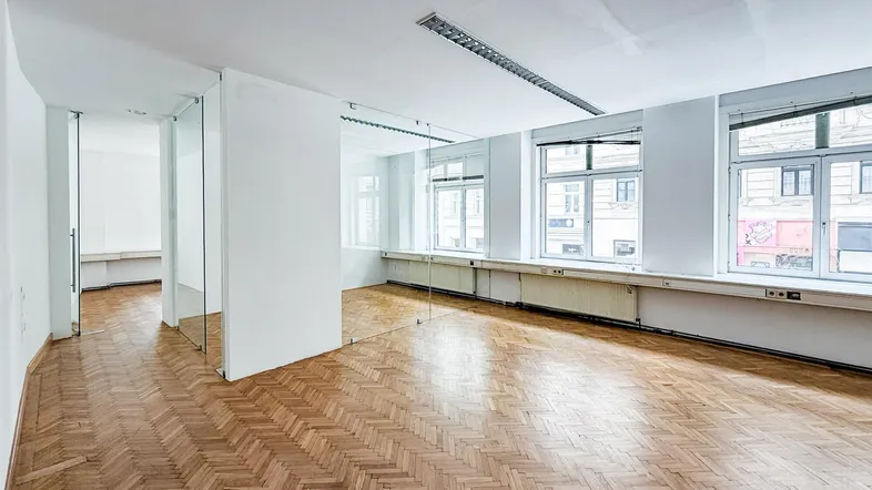 Geräumiges Büro mit Fischgrätparkett, großen Fenstern und modernen Glastrennwänden für eine offene Arbeitsumgebung.