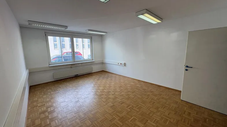 Leerer Büroraum mit Parkettboden, großen Fenstern und Deckenleuchten, bereit zur individuellen Gestaltung.