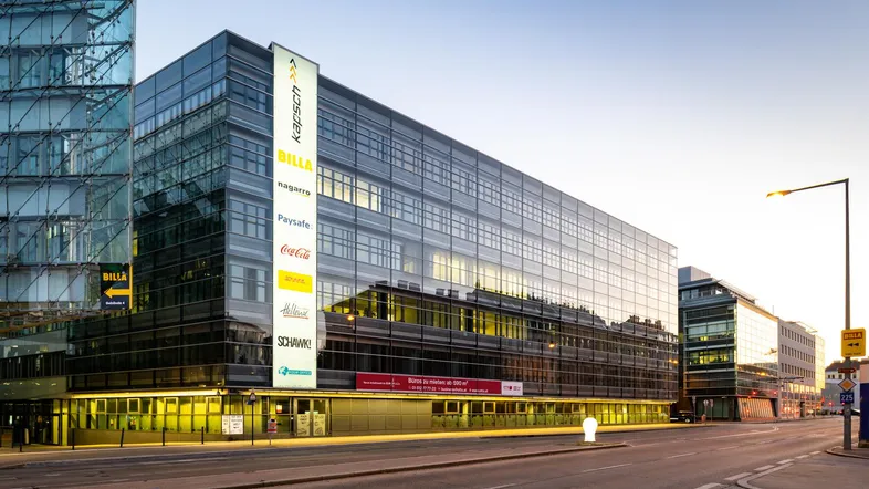 Modernes Bürogebäude mit Glasfassade und Werbetafeln an einer belebten Straße bei Sonnenuntergang.