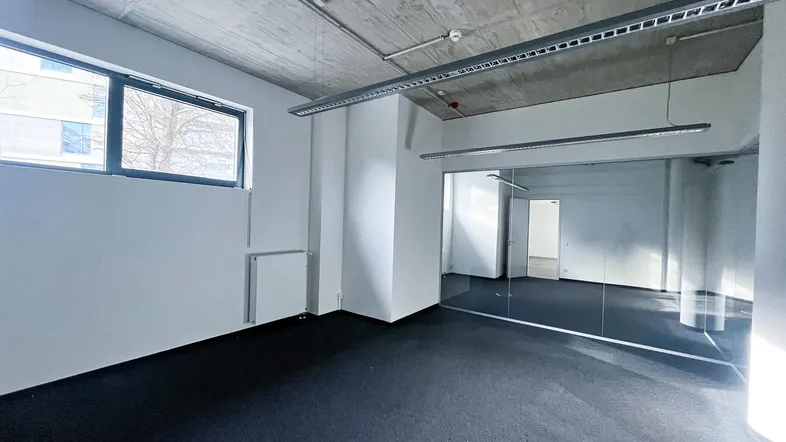Geräumiges Büro mit dunklem Teppichboden, großen Fenstern und sichtbarer Betondecke, ideal für moderne Arbeitsumgebungen.