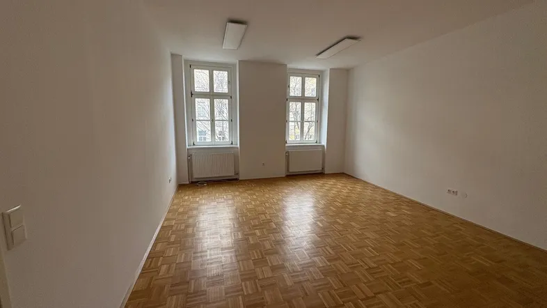 Heller Raum mit Fischgrätparkett, weißen Wänden und zwei Fenstern, ideal als Büro oder Wohnbereich.