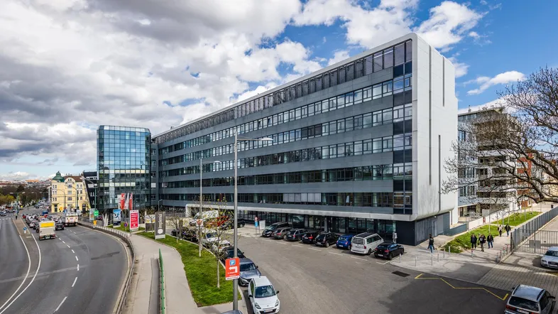 Modernes Bürogebäude mit Glasfassade und Parkplätzen an einer belebten Straße unter bewölktem Himmel.