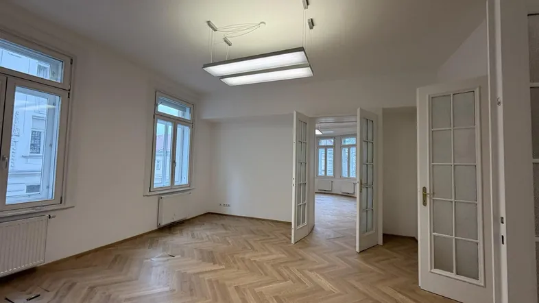 Geräumiger, leerer Raum mit Fischgrätparkett und großen Fenstern, der viel natürliches Licht hereinlässt.