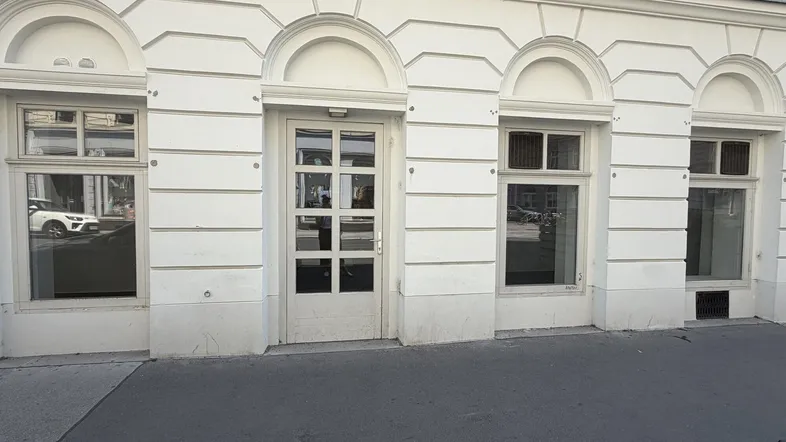 Die weiße Fassade des Erdgeschosses zeigt eine Eingangstür und mehrere Fenster zur Straße hin.