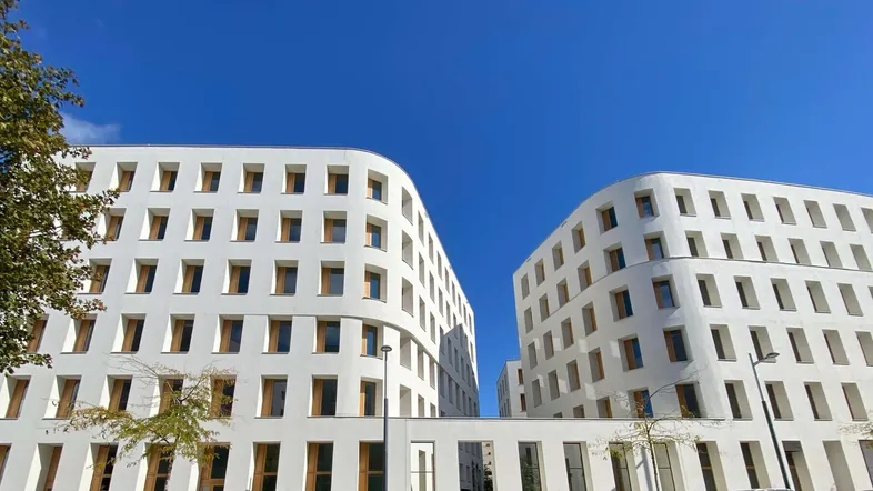 Zwei moderne, weiße Gebäude mit vielen kleinen Fenstern unter blauem Himmel und einem Baum.