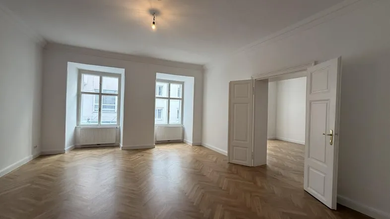 Geräumiges Wohnzimmer mit klassischem Fischgrätparkett, hohen Decken und zwei großen Fenstern, die viel Tageslicht hereinlassen.