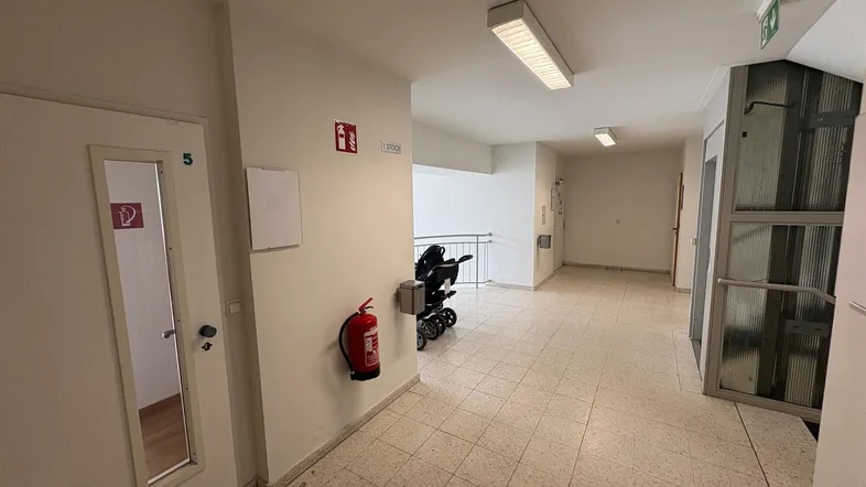 Geräumiger Flur mit Fliesenboden, Feuerlöscher und Zugang zu einem Aufzug und Treppenhaus.