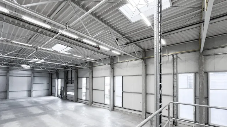 Geräumige, leere Industriehalle mit hohen Decken, Betonboden und viel Tageslicht durch Oberlichter und Fenster.