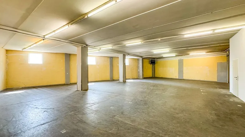 Großer, leerer Lagerraum mit gelben Wänden, Betonboden und sichtbaren Stützpfeilern, beleuchtet von Deckenleuchten.