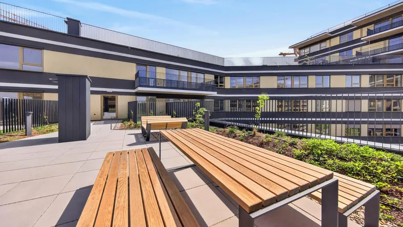 Moderne Dachterrasse mit Holzbänken und Tischen, umgeben von zeitgenössischen Gebäuden und Grünflächen.