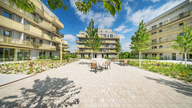 Moderne Wohngebäude umgeben einen gepflasterten Innenhof mit Sitzgelegenheiten und jungen Bäumen.