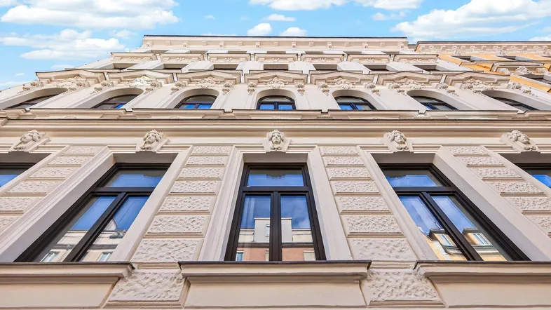 Historische Fassade eines Altbaus mit reich verzierten Fenstern und Stuckelementen unter blauem Himmel.