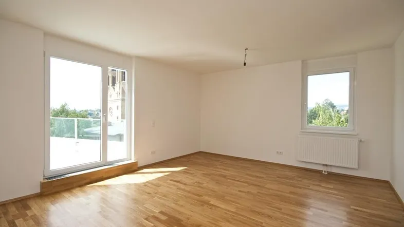 Helles Zimmer mit Parkettboden, großen Fenstern und Zugang zu einem Außenbereich mit Blick auf eine Kirche.