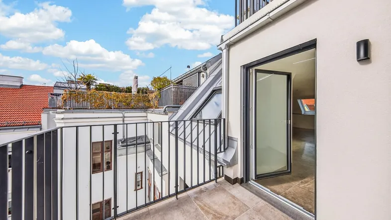 Heller Balkon mit Fliesenboden und Blick auf die umliegenden Dächer und den blauen Himmel.