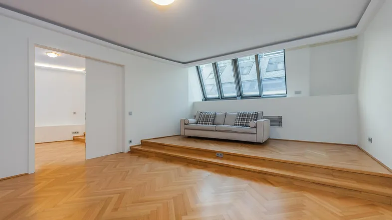 Helles Wohnzimmer mit erhöhtem Podestbereich, großen Fenstern und Fischgrätparkettboden.