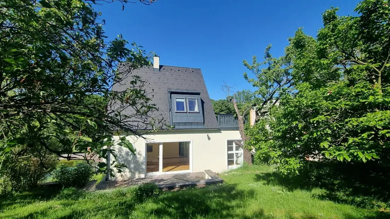 Hausfassade mit großen Glasfronten und Zugang zum grünen Garten unter blauem Himmel.