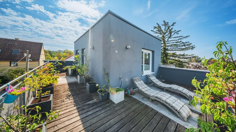 Geräumige Dachterrasse mit Holzbelag, Liegestühlen und Pflanzen unter blauem Himmel.