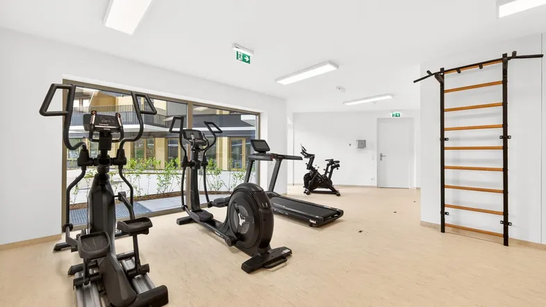 Moderner Fitnessraum mit verschiedenen Trainingsgeräten und großen Fenstern für natürliches Licht.