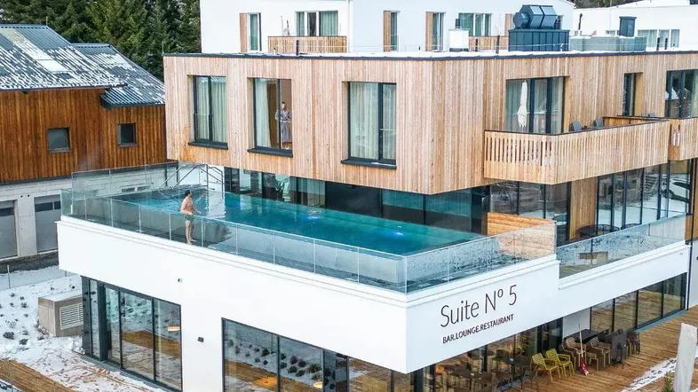 Modernes Gebäude mit Holzfassade und einem beeindruckenden Infinity-Pool auf der Dachterrasse, umgeben von verschneiten Bäumen.