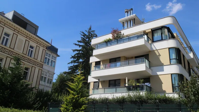 Modernes Mehrfamilienhaus mit geschwungenen Balkonen und einer traditionellen Villa im Hintergrund.
