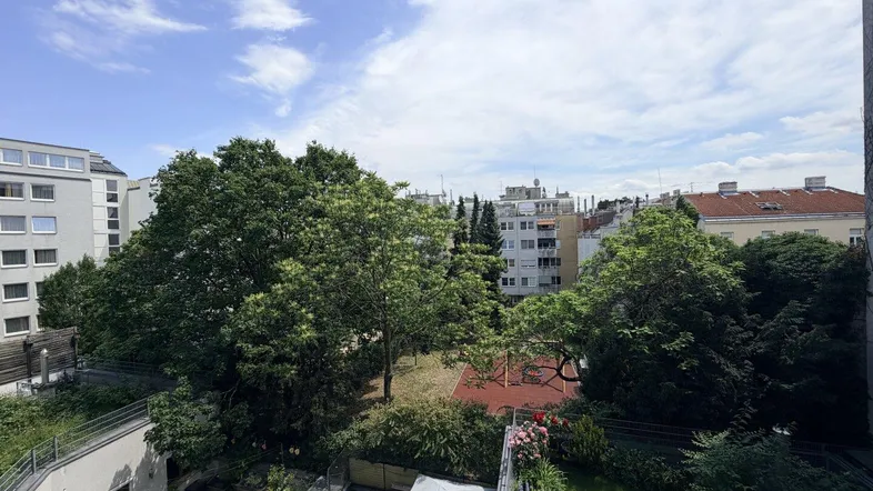 Blick von oben auf einen grünen Innenhof mit Bäumen, Spielplatz und umliegenden Gebäuden unter blauem Himmel.