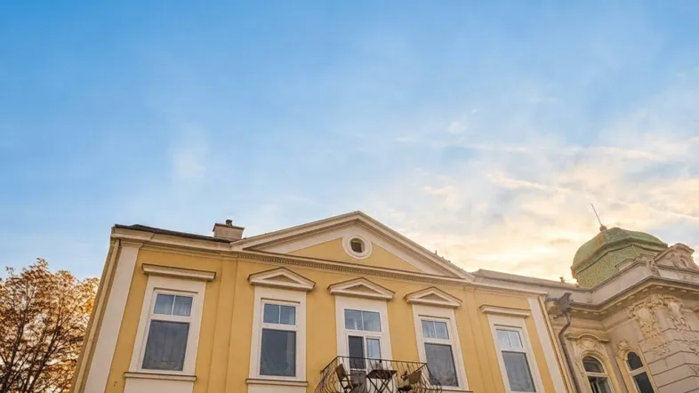 Gelbe Fassade eines historischen Gebäudes mit Balkon und grüner Kuppel unter blauem Himmel.